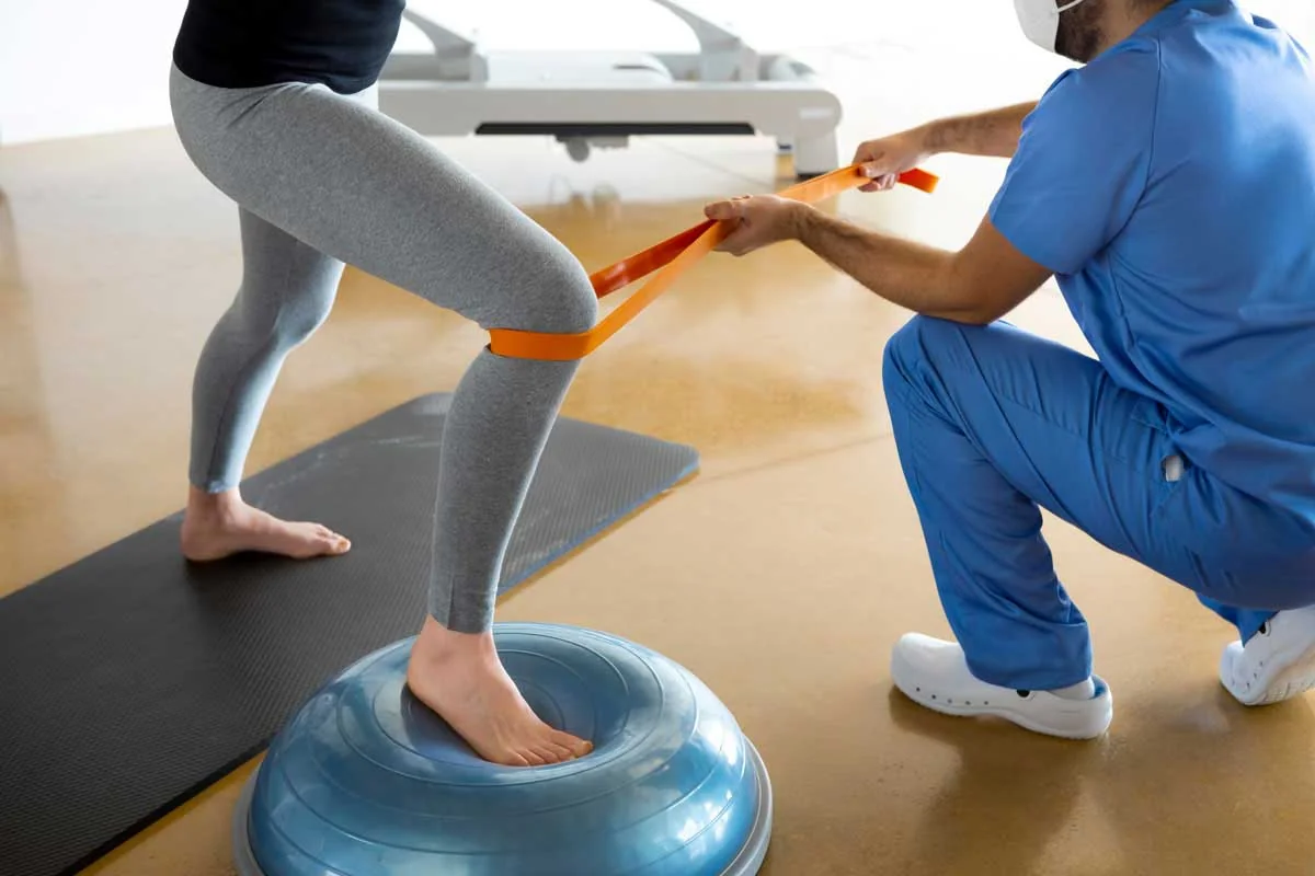 Krankengymnastik am Gerät: Patient trainiert mit Widerstandsband auf Balance-Plattform unter therapeutischer Anleitung bei PHYSIOEFFEKT Berlin
